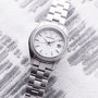 The Grand Seiko Ladies Heritage wristwatch features a silver case, snowflake dial, date display, blued steel seconds hand, and 44 diamonds on the bezel, displayed on a textured white and gray surface.