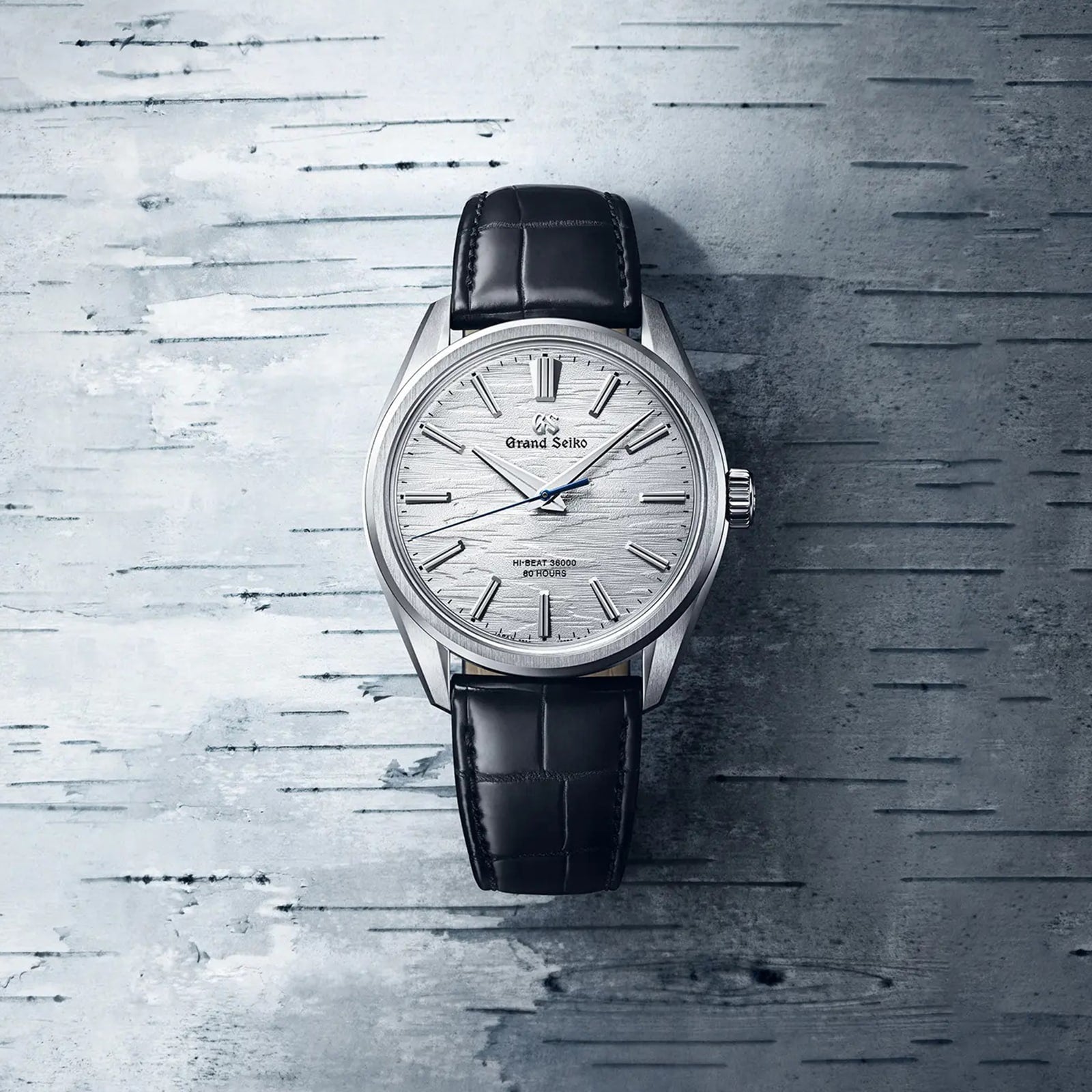 A Grand Seiko Manual Hi-beat 80 Hours Evolution 9 wristwatch with a textured white birch dial and black leather strap, shown on a gray patterned surface.