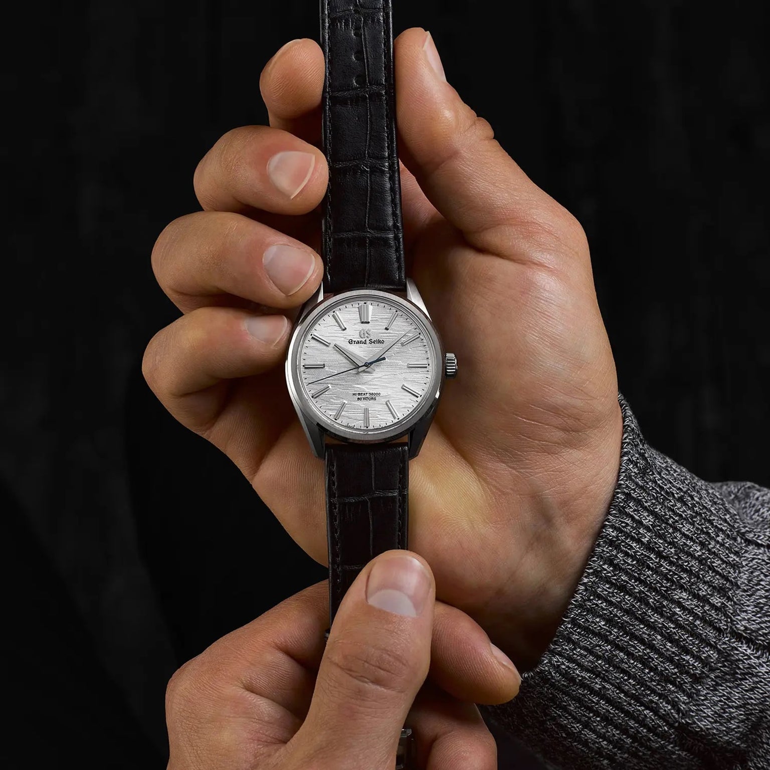 A person holds a Grand Seiko Manual Hi-beat 80 Hours Evolution 9 wristwatch featuring a textured white birch dial, silver case, and black leather strap against a black background.