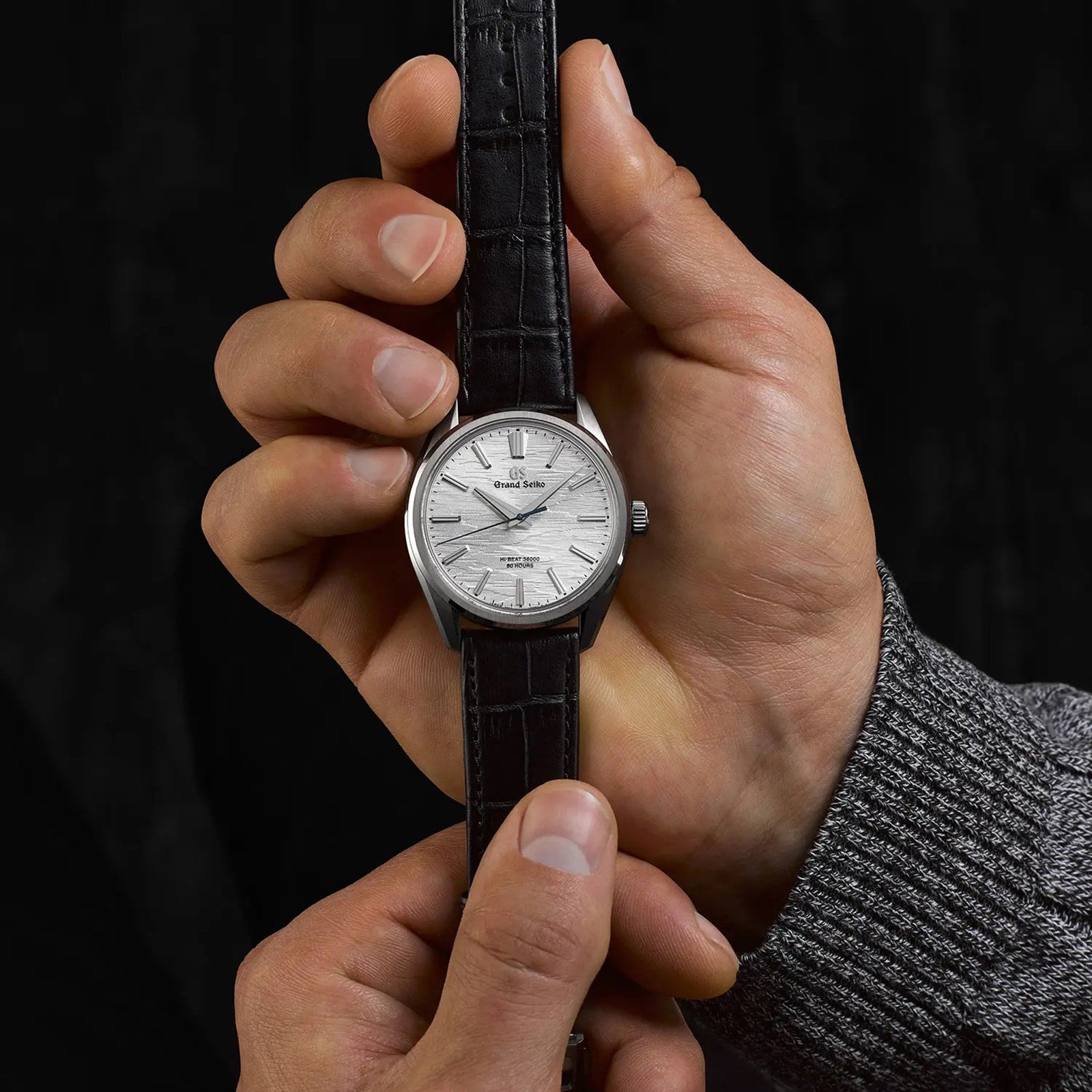 A person holds a Grand Seiko Manual Hi-beat 80 Hours Evolution 9 wristwatch featuring a textured white birch dial, silver case, and black leather strap against a black background.