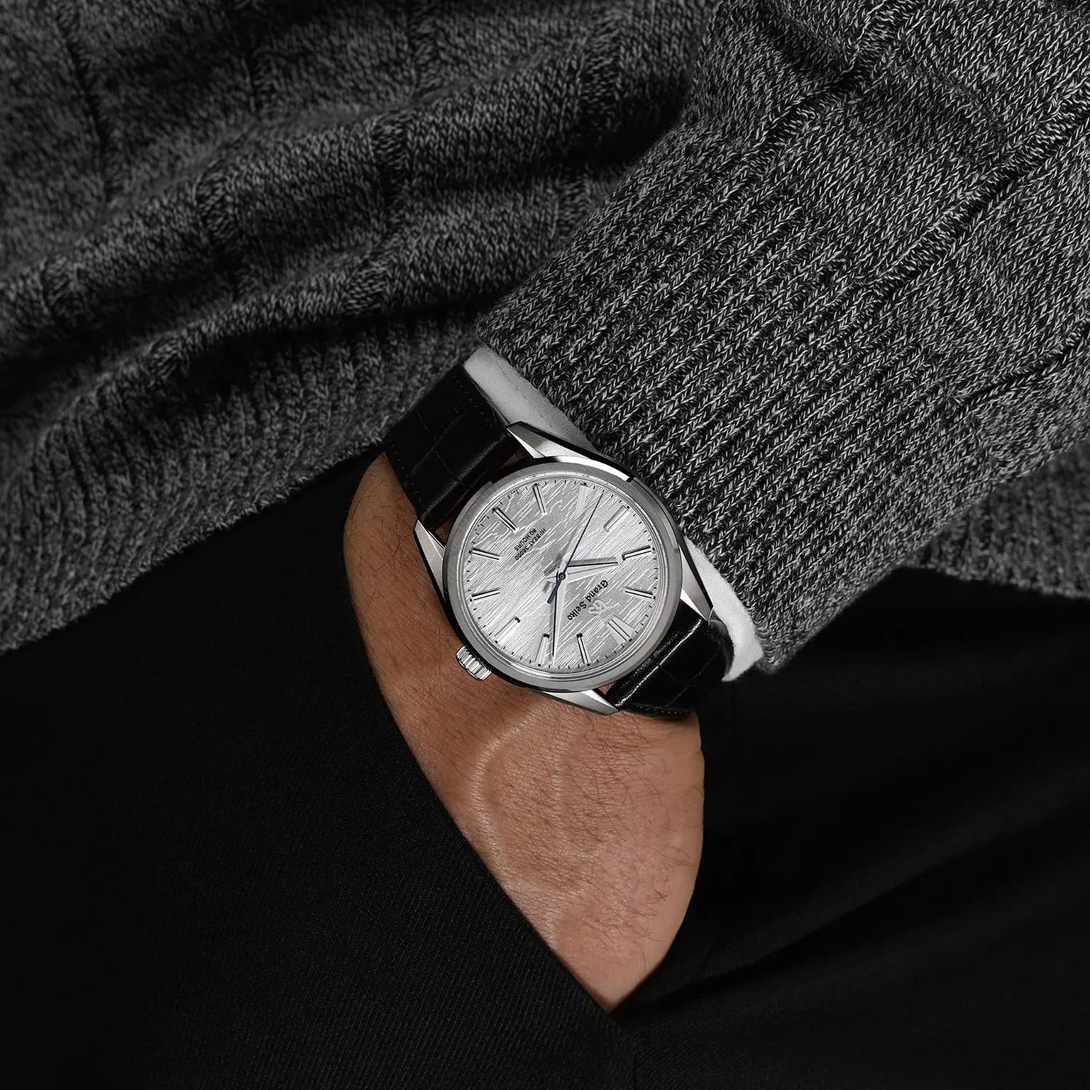 A person in a gray sweater and black pants has their hand in their pocket, showcasing the Grand Seiko Manual Hi-beat 80 Hours Evolution 9 wristwatch with a silver dial and black leather strap.