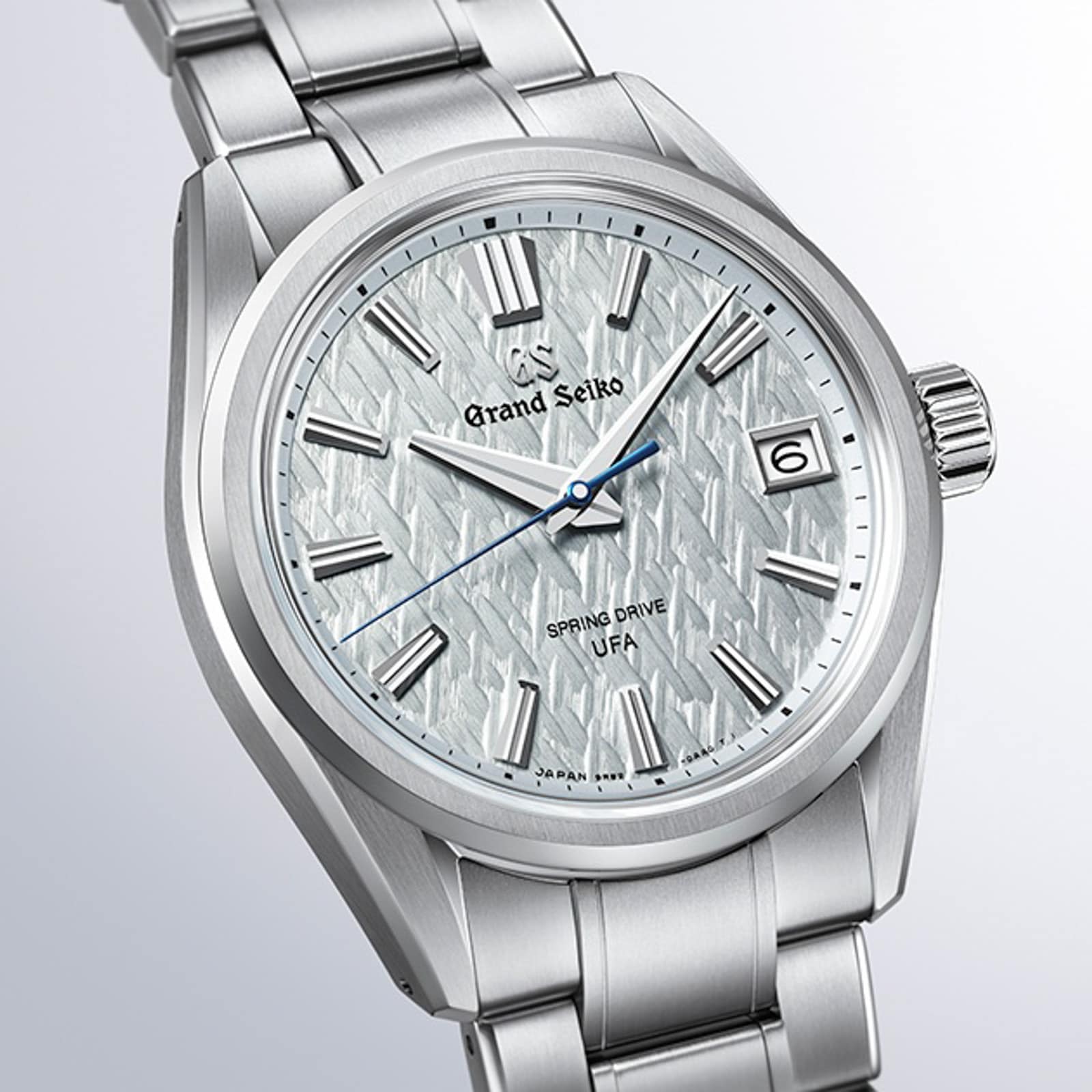 The Grand Seiko Spring Drive U.F.A Evolution 9 wristwatch features a silver case, textured white dial, date window, and blue second hand.
