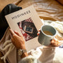 A person holds HODINKEE Magazine, Volume 9 by Hodinkee and a gray mug, wearing a wristwatch—ideal for horology lovers—while relaxing on a tan leather sofa with a blanket.