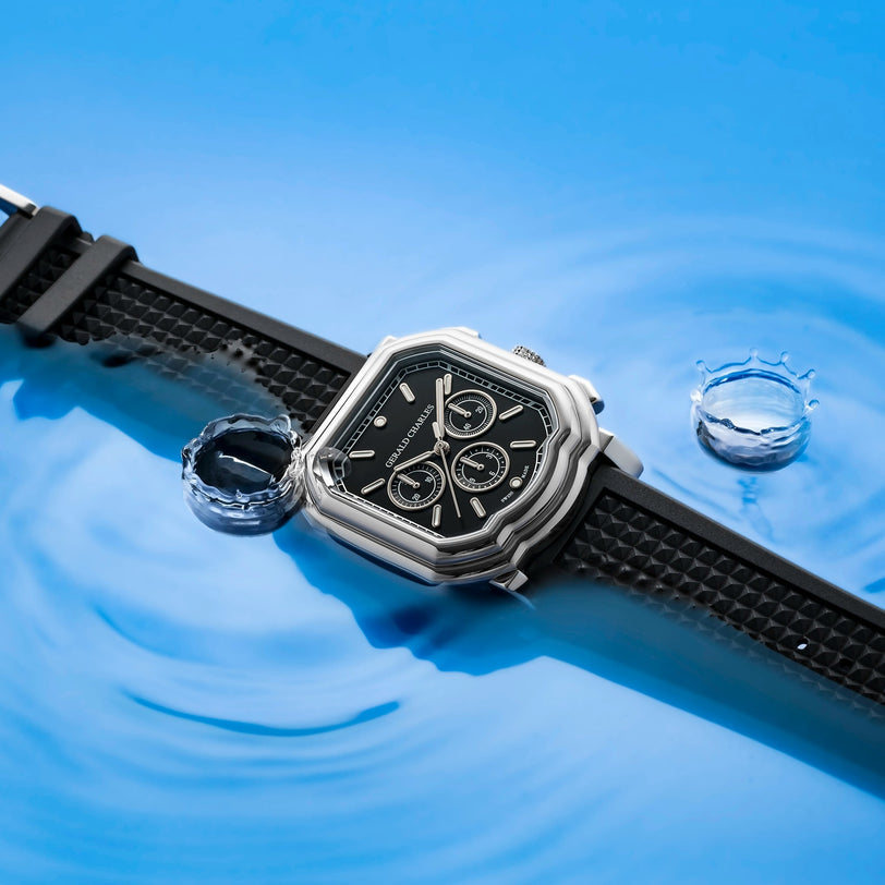 The Gerald Charles Maestro 3.0 Chronograph, a stainless steel watch with a black strap, is showcased on a blue surface with water ripples and splashes, emphasizing its water-resistant design.