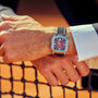 Close-up of a person wearing the Gerald Charles Maestro GC Sport, a maroon watch with a black strap, standing by a sports net.