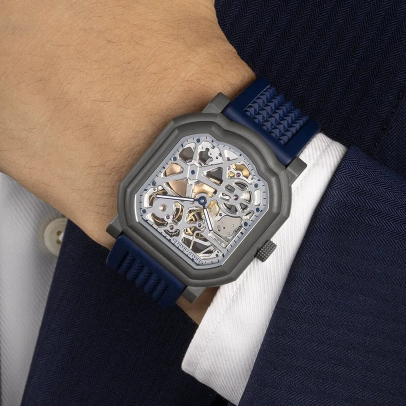 A person in a navy suit wears the Gerald Charles Maestro 8.0 GC Sport Squelette, a square skeleton watch with a blue strap, on their wrist.