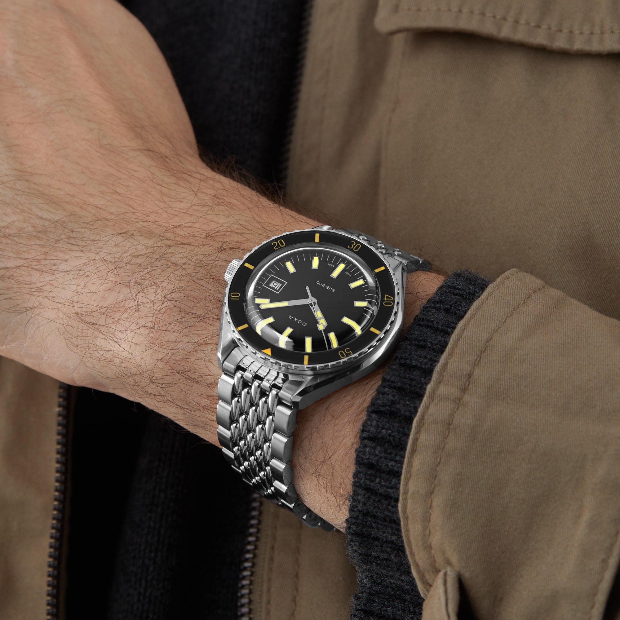 A person wearing the DOXA Sub 200 Sharkhunter divers watch, featuring a black and yellow dial with a unidirectional rotating bezel, paired with a brown jacket and black sweater.