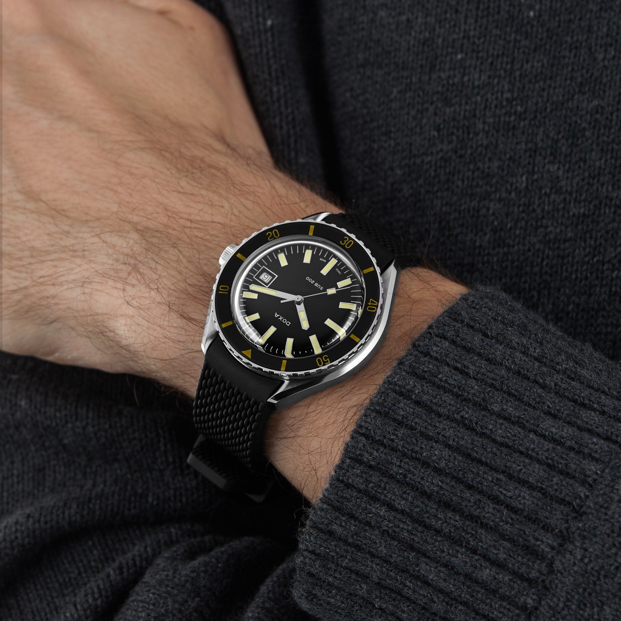 A person wearing a DOXA Sub 200 Sharkhunter with a 316L stainless steel mesh strap on their left wrist, dressed in a dark knit sweater.