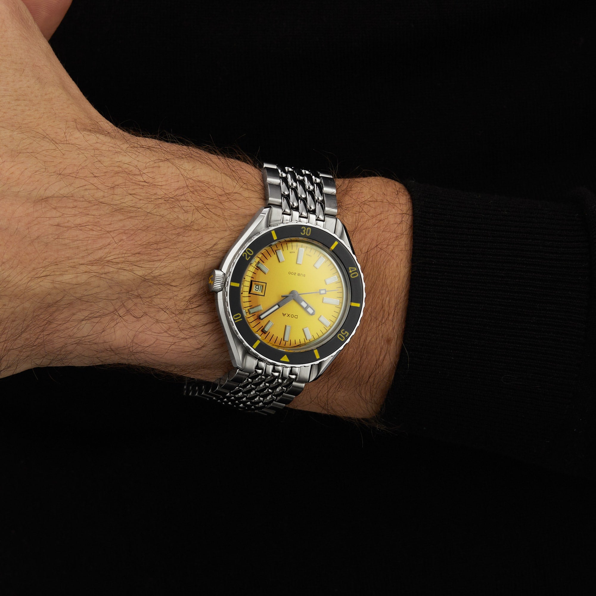 A person wearing a DOXA Sub 200 Divingstar with a yellow face and metal band on their left wrist, dressed in a long-sleeved black shirt.