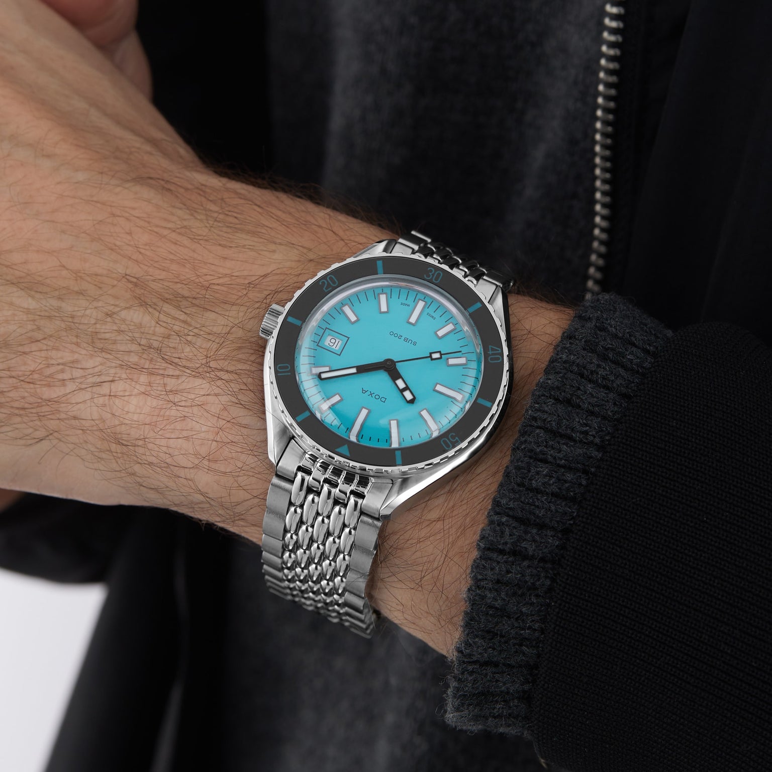 A person wearing the DOXA Sub 200 Aquamarine, a divers watch featuring a bright turquoise face and silver 316L stainless steel band, shown on their left wrist with a dark jacket and grey sweater.