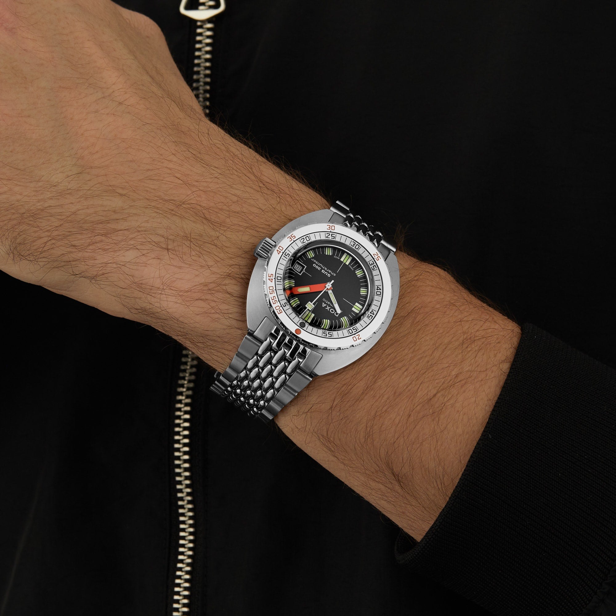Wearing a DOXA Sub 300 Sharkhunter—silver divers watch with black dial and green accents, unidirectional bezel—a person stands against a black background in a black zippered jacket.