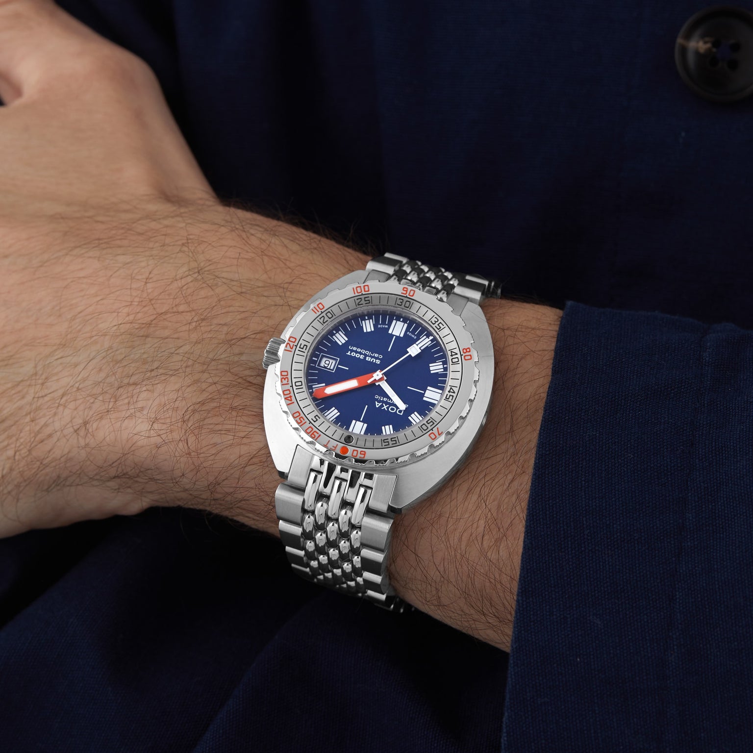 The DOXA Sub 300T Caribbean, featuring a stainless steel band and blue dial, is worn on a persons left wrist against a dark blue sleeve.