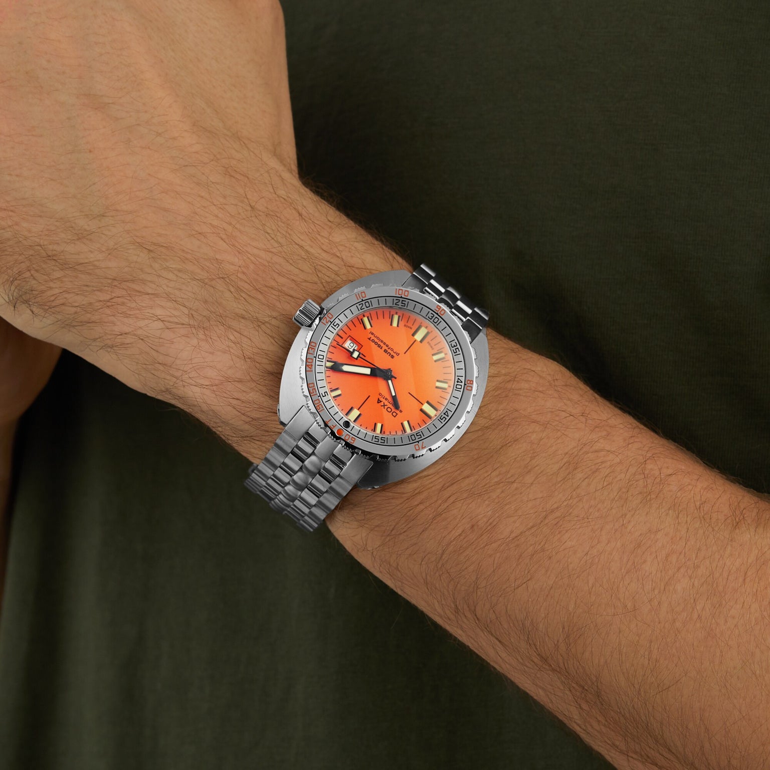 A person wears a DOXA Sub 1500T Professional, a stainless steel divers watch with an orange dial and metal bracelet, on their left wrist against a green shirt background.