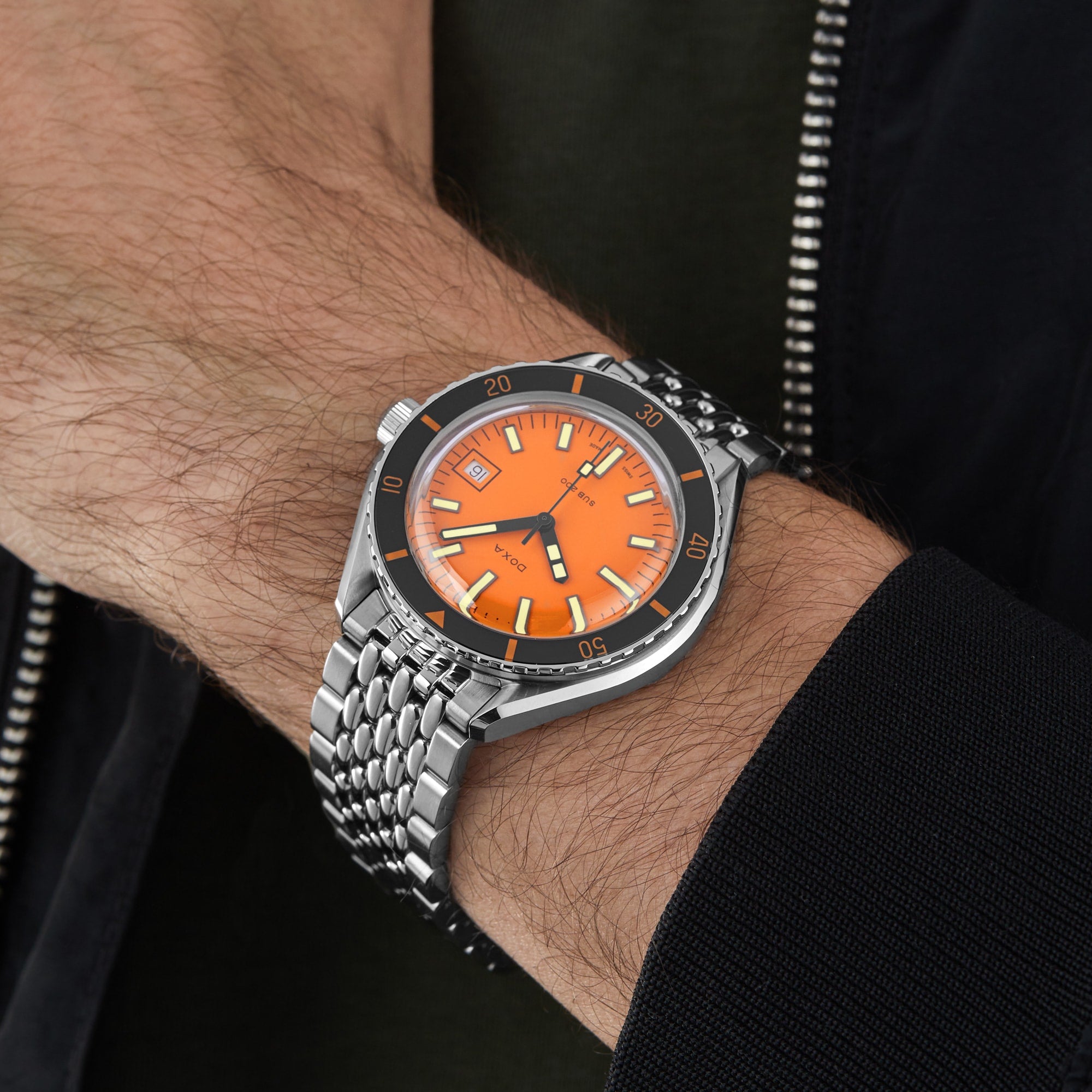 A person wearing the DOXA Sub 200 Professional diver’s watch with an orange dial, 316L stainless steel band, and unidirectional rotating bezel on their left wrist, dressed in a dark jacket and green shirt.