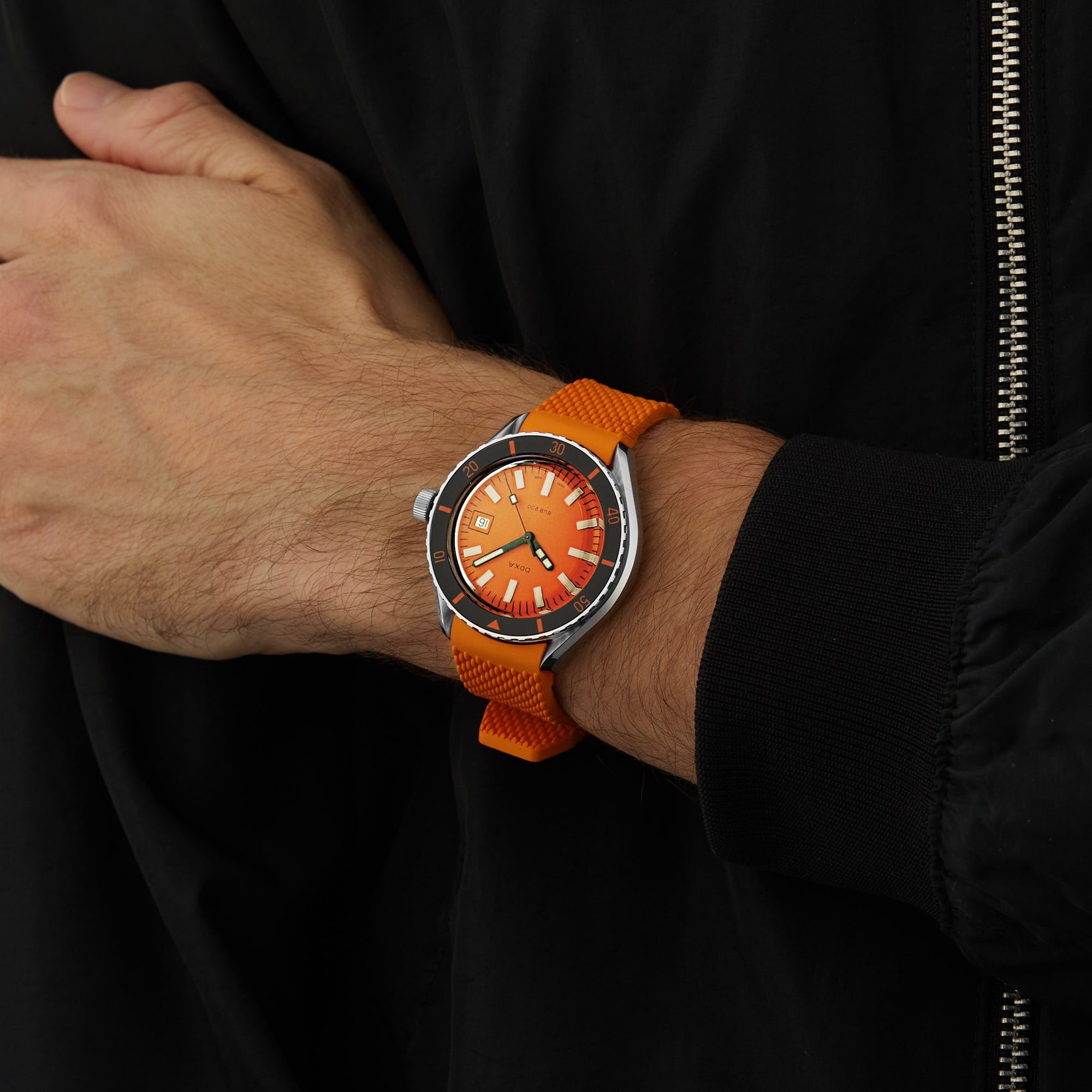 A person wearing the DOXA Sub 200 Professional divers watch with an orange strap and dial, featuring a unidirectional rotating bezel, is dressed in a black jacket with a visible zipper.