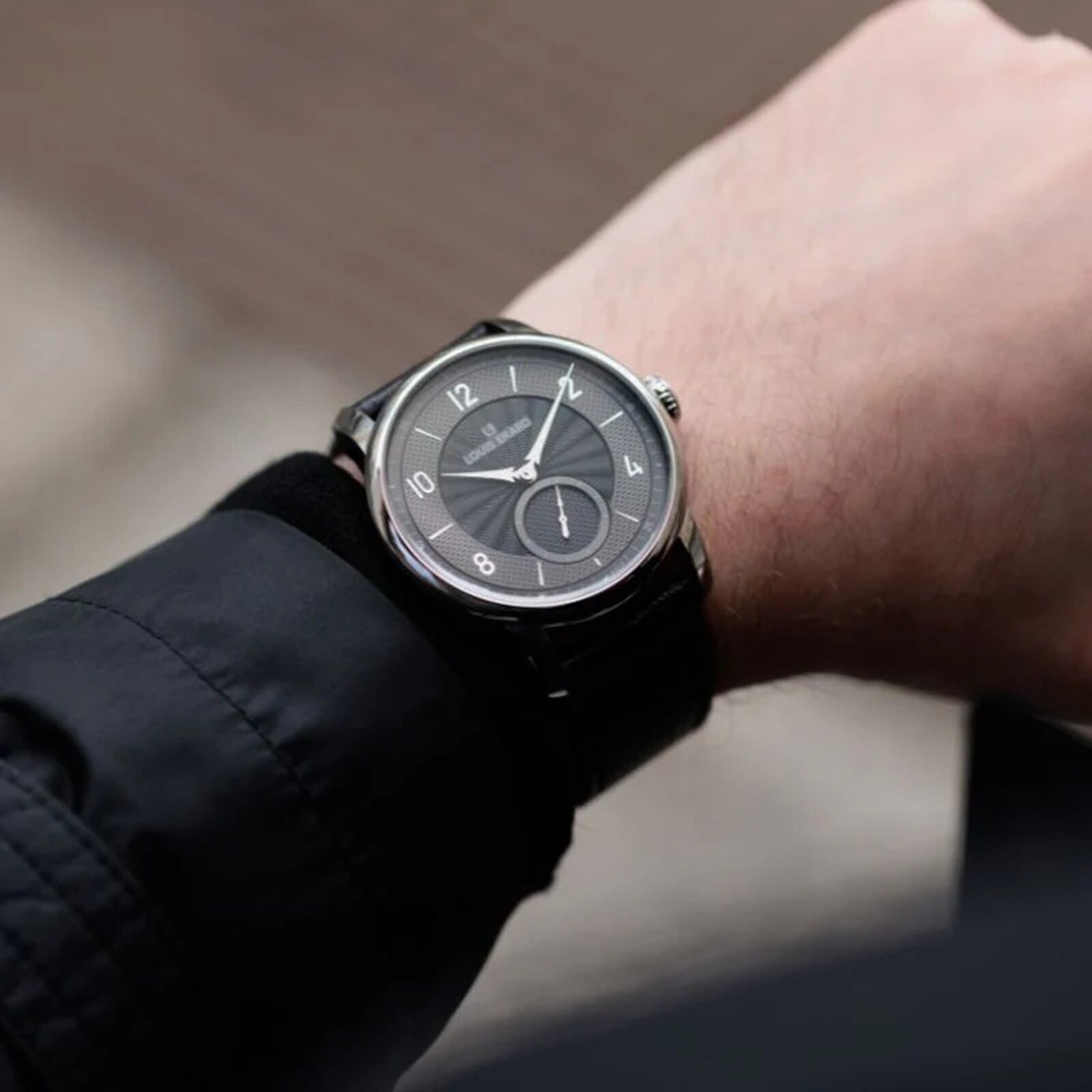 A person wearing a Louis Erard Petite Seconde Guilloché Anthracite Black wristwatch with a dark dial and silver hour markers on their left wrist.