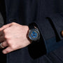 A person wearing a dark coat and a Louis Erard Petite Seconde Guilloché Anthracite Blue watch with a blue face and black strap on their left wrist.