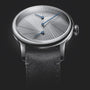 The Louis Erard Le Régulateur, created with atelier oï, features a silver dial with a radiating texture, blue hands, and a black leather strap, showcased against a dark backdrop.