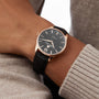 A person wearing the William Wood Chivalrous Collection - Rose Gold watch, featuring a black leather strap, black face, and elegant rose gold casing.