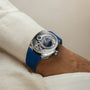 The Greubel Forsey GMT Balancier Convexe, a silver luxury watch with a blue strap, is worn on a wrist in a cream-colored sleeve.