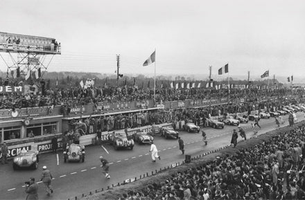 Drivers sprint to their cars at the start of a historic Le Mans race, with crowds watching from packed grandstands.