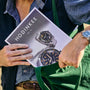 A person, wearing a watch on their left wrist, slides the HODINKEE Magazine, Volume 12 by Hodinkee—featuring watches on the cover—partially into a green bag, capturing vintage watch style.