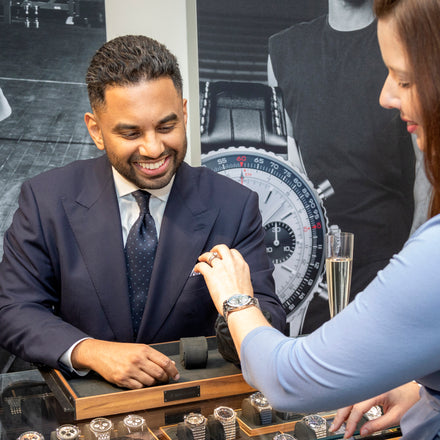 A smiling man in a suit assists a woman trying on a watch at a jewelry store counter.