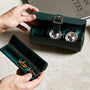 Hands holding the WOLF British Racing Triple Watch Roll in green leather with two watches, alongside another case containing gold cufflinks, all placed on a stone surface.