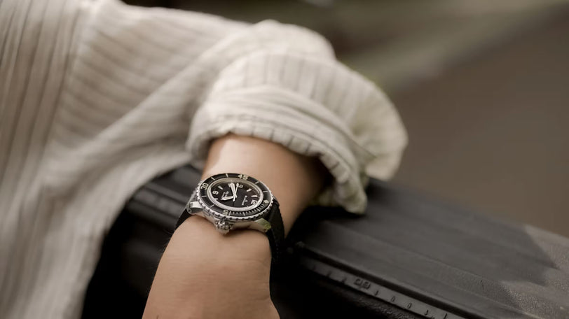 Watch with black sunburst dial and rubber strap worn on wrist, featuring round case with tachymeter bezel.