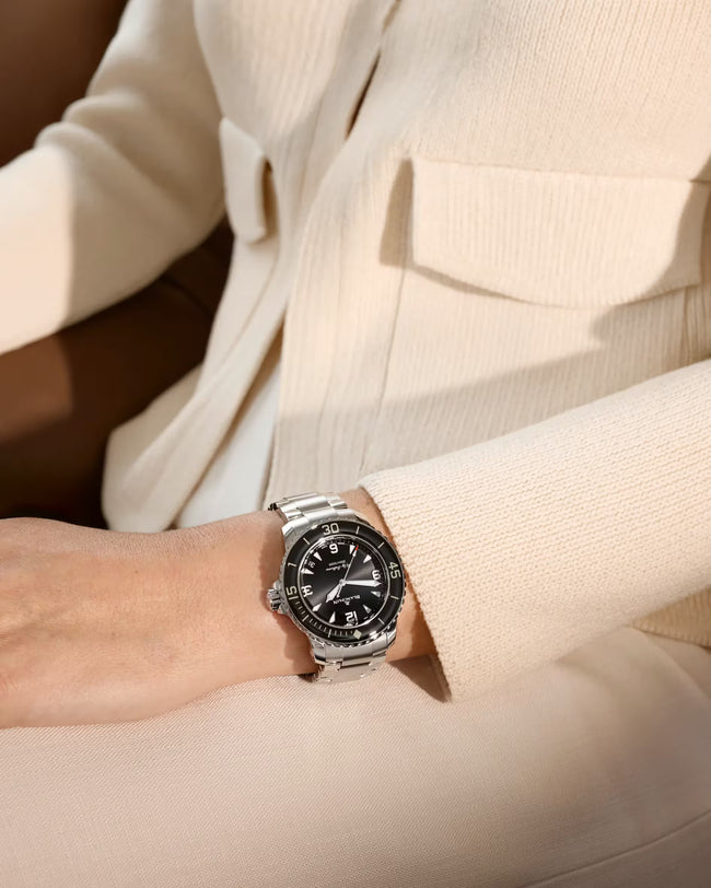 Fifty Fathoms watch with sunburst black dial and steel bracelet worn on wrist against cream-colored fabric.