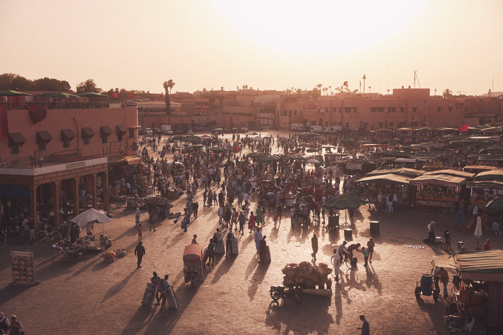 A bustling outdoor market square at sunset, filled with people, stalls, and warm golden light.