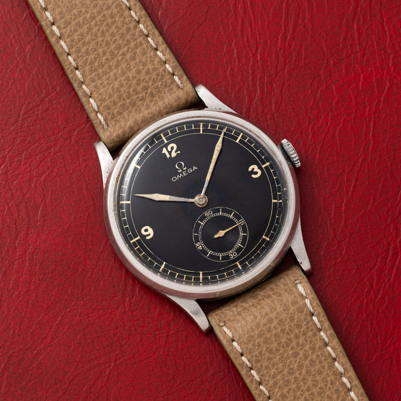 An OMEGA Jumbo Calatrava wristwatch with a black dial and tan leather strap displayed on a red textured surface.