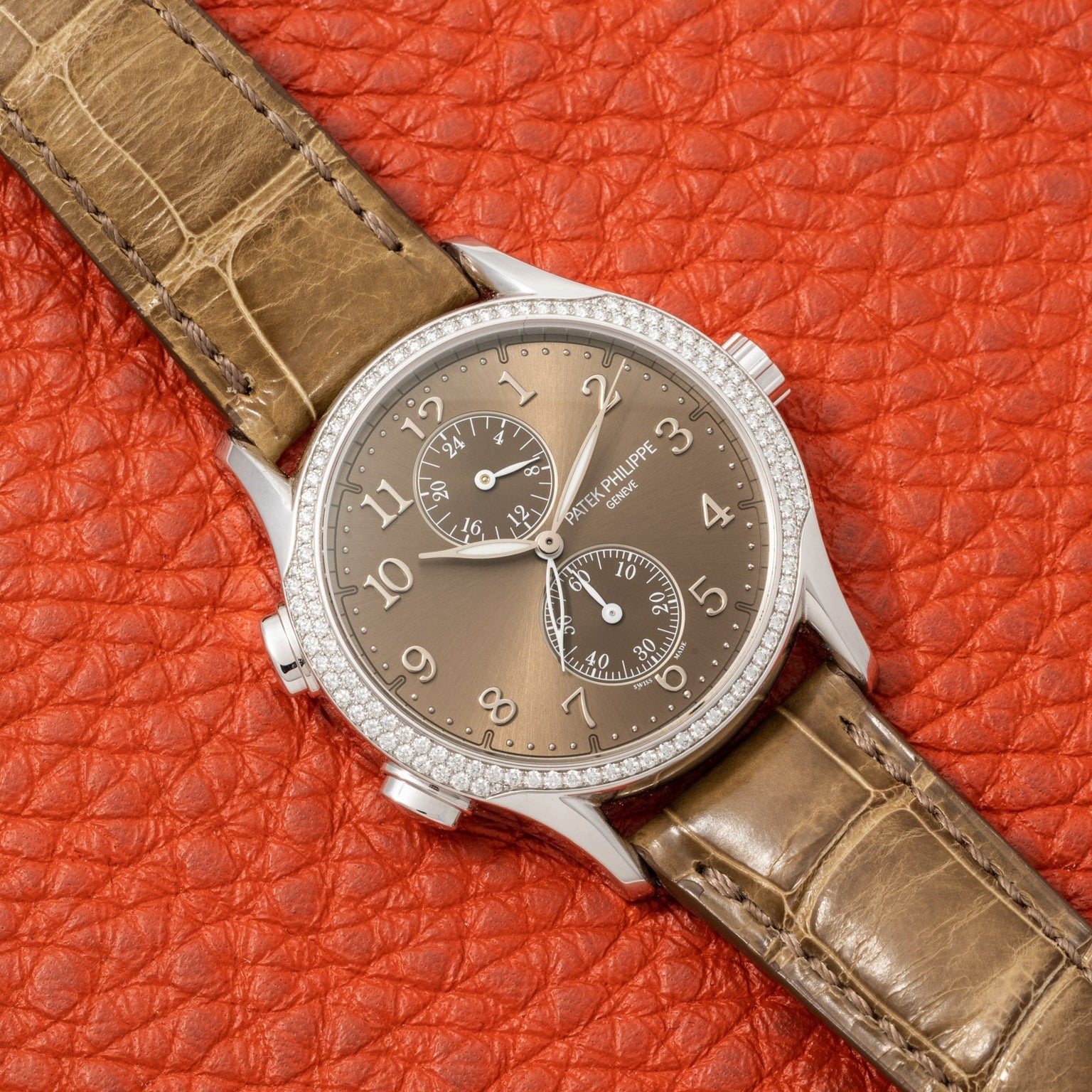 A Patek Philippe Calatrava Travel Time Ladies watch featuring a diamond bezel and leather strap is displayed on textured orange leather.