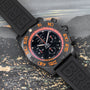 The Breitling Chronomat Raven chronograph features orange accents on a black rubber strap, showcased against a cityscape background.