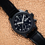 The IWC Schaffhausen Pilots Watch Chronograph Tribute To 3705 with a black dial and leather strap is displayed on a textured beige surface.