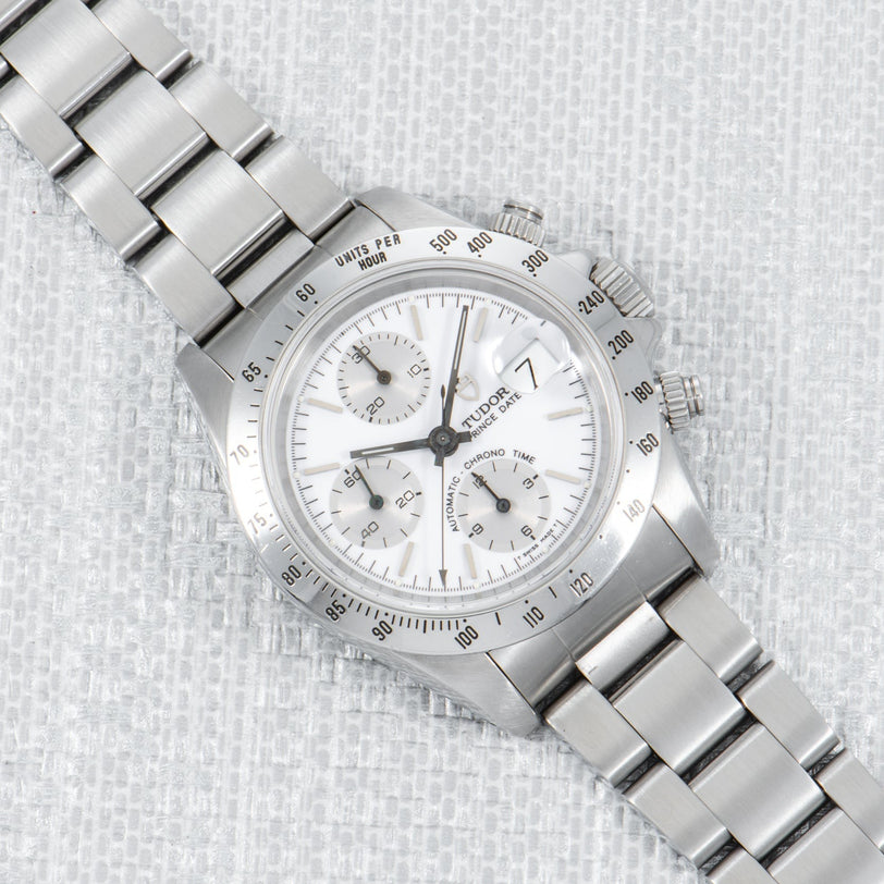 A TUDOR Prince Date Chronograph Silver Albino wristwatch with a stainless steel case, white dial, and metal bracelet, displayed on textured fabric.