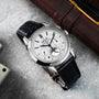 The Patek Philippe Perpetual Calendar Moonphase Chronograph is a luxury wristwatch featuring a black leather strap, white dial, multiple subdials, and date indicators.