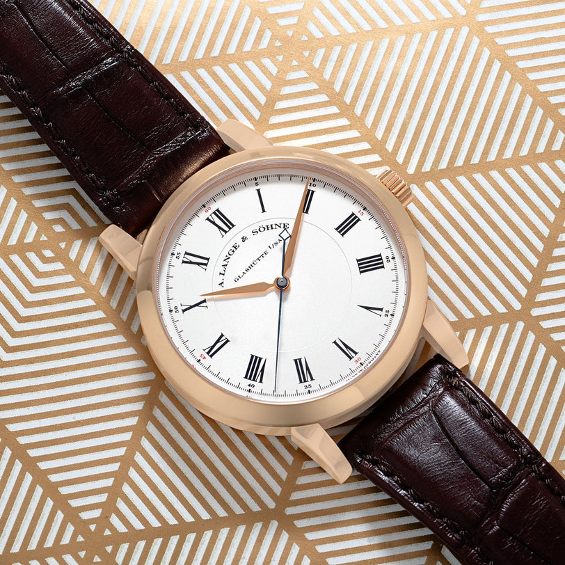 The Richard Lange by Lange & Sohne features a white face, black Roman numerals, and a brown leather strap, displayed on a geometric gold background.
