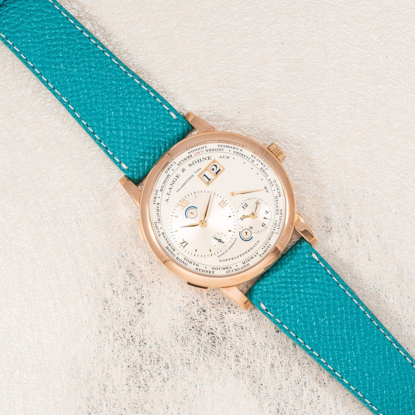The Lange & Sohne Lange 1 Timezone gold wristwatch features a white dial and a bright turquoise leather strap, displayed on a light textured surface.