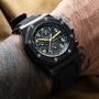 The Audemars Piguet Royal Oak Offshore End of Days chronograph is shown on a tattooed wrist, its yellow hands contrasting against the black watch and a brown fabric background.
