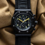 The Audemars Piguet Royal Oak Offshore End of Days showcases yellow hands on a black chronograph watch set against a textured tan fabric background.