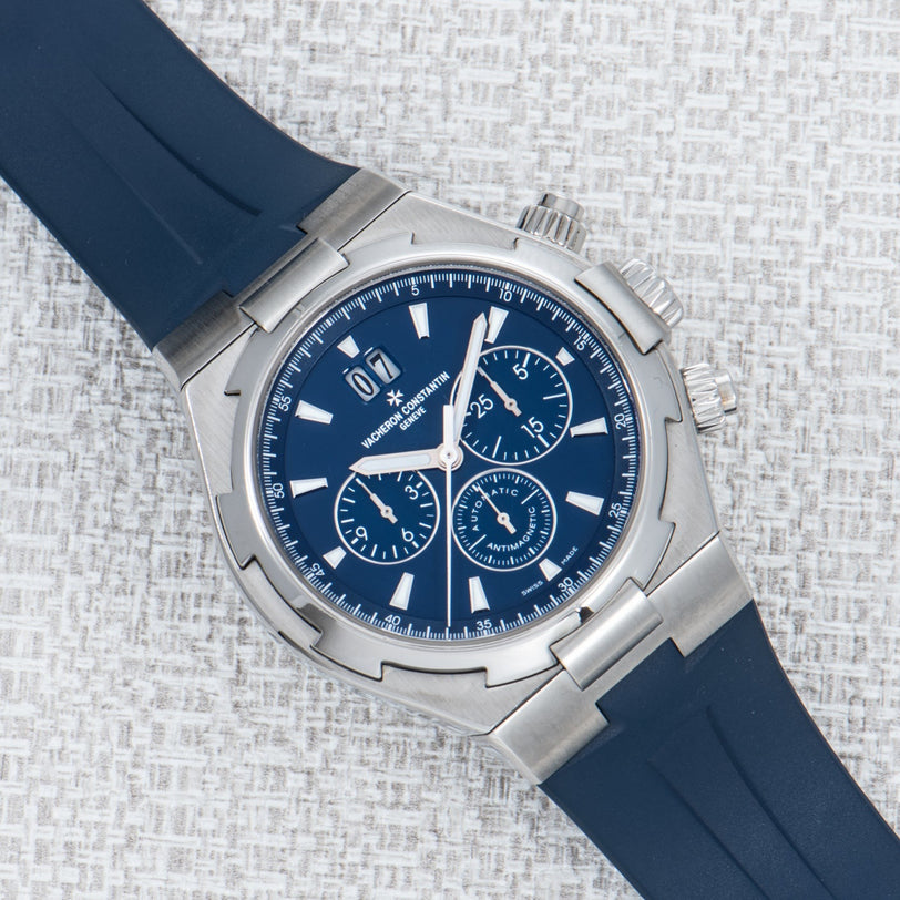 The Vacheron Constantin Overseas Chronograph is a luxury silver watch with a blue face and blue rubber strap, shown on a textured light surface.