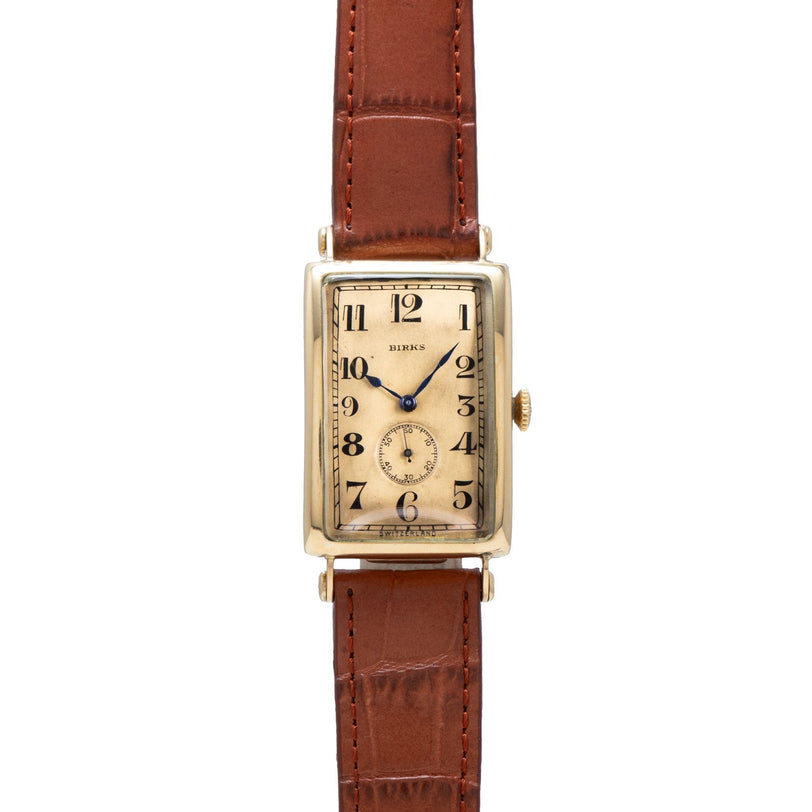 The Patek Philippe Birks Rectangular Dress Watch features a gold case, brown leather strap, and black numerals on a beige face.
