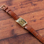 A Patek Philippe Birks Rectangular Dress Watch with a gold case and brown leather strap displayed on a wooden surface.