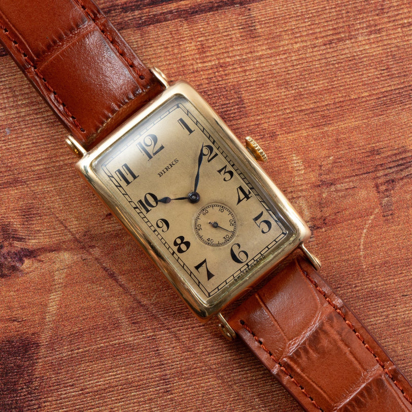 Patek Philippe Birks Rectangular Dress Watch featuring a gold case and brown leather strap displayed on a textured brown surface.