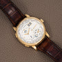 The Lange & Sohne Lange 1 Timezone is a gold wristwatch with a brown leather strap, large date display, multiple subdials, and a textured dial.
