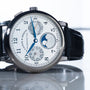 The Lange & Sohne Lange 1815 Annual Calendar, featuring a black leather strap and multiple subdials, is displayed on a reflective surface.