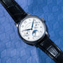 Lange & Sohnes Lange 1815 Annual Calendar analog wristwatch with a black leather strap displayed on a textured blue surface.