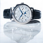 The Lange & Sohne Lange 1815 Annual Calendar wristwatch, featuring a black leather band and multiple dials, is reflected on a shiny surface.