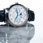 The Lange & Sohne Lange 1815 Annual Calendar features blue hands, a black leather strap, and multiple subdials, all elegantly reflected on a shiny surface.